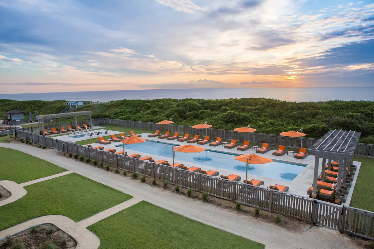 A vibrant exterior shot focusing on the resort's swimming pool surrounded by orange umbrellas and lounge chairs. In the distance, the sun sets over the ocean, casting a warm glow across the space. The image conveys relaxation, social opportunities, and the proximity of the balcony spaces to both the pool and the beach.