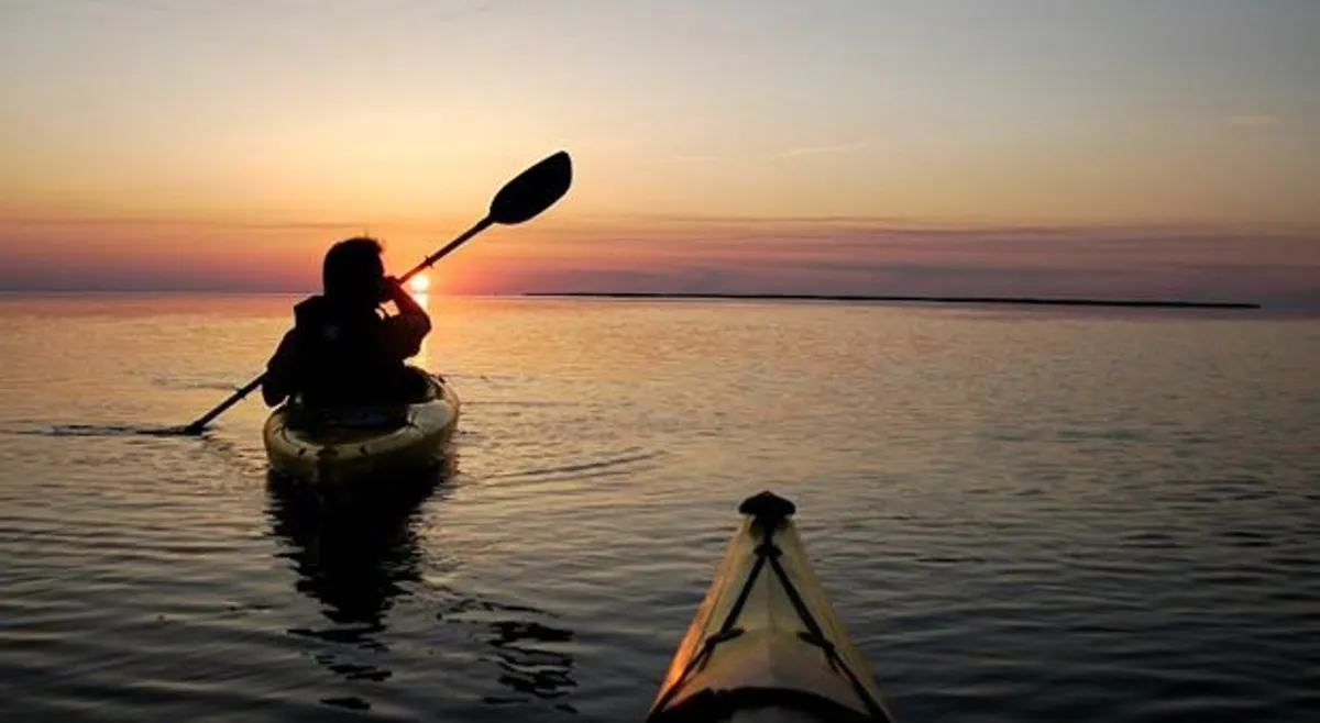 A single kayaker drifts peacefully on calm water at sunset, with the golden sky reflecting across the sound. The tranquil setting highlights the natural beauty and serenity of waterfront moments, ideal for intimate or reflective experiences.