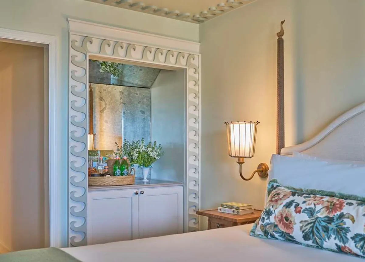 This interior photo highlights a serene corner of the guestroom, featuring a decorative mirror, a floral pillow on a plush bed or chair, a wall sconce providing gentle light, and a lush green plant arrangement. The natural light streaming in suggests an inviting and peaceful atmosphere for relaxation or reading.