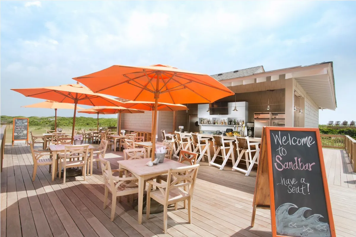 A lively outdoor dining area shaded by bright orange umbrellas, with wooden tables and a visible bar area. Guests can enjoy meals, drinks, and relaxed conversations while taking in ocean views. The casual, breezy mood is perfect for lunches, cocktails, or sunset gatherings.