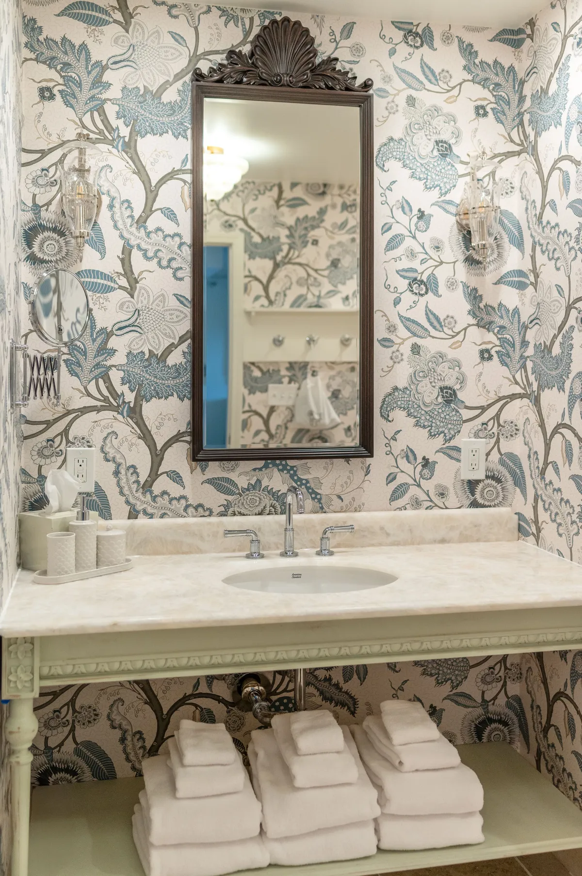 A beautifully styled ensuite bathroom with an ornate mirror, striking floral wallpaper, a marble countertop with an elegant sink, and neatly stacked towels. The scene evokes a spa-like, calming vibe.