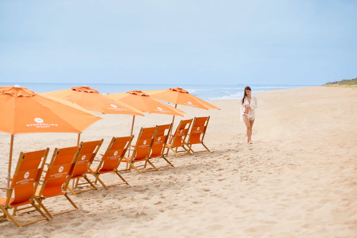 A serene beach scene with clusters of beach chairs and umbrellas facing the ocean, a woman strolling along the shore, and inviting spots for families to relax or build sandcastles. The tranquil setting highlights relaxation, sand play, and scenic ocean views.