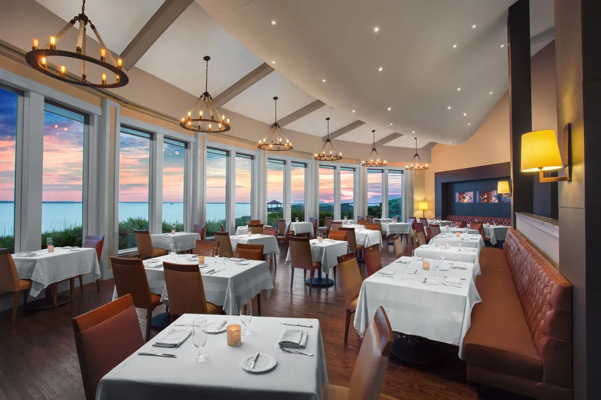 An elegantly set interior restaurant space, with large sunset-lit windows, beautifully arranged tables, and sparkling chandeliers. The ambiance is sophisticated yet warm, perfect for romantic dinners or special occasions. The setting emphasizes both fine cuisine and memorable experiences.