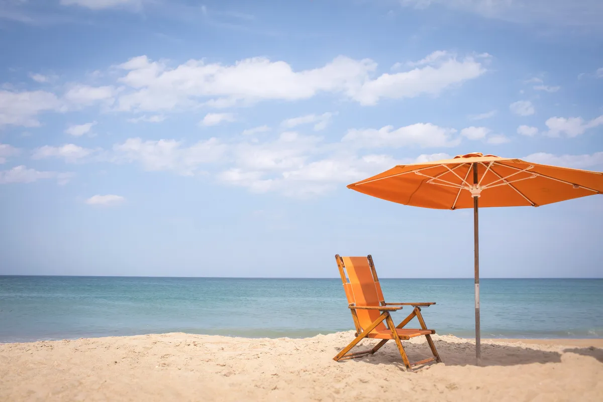 A serene stretch of sandy beach featuring an inviting orange beach chair and matching umbrella set up close to the calm sea. The scene exudes relaxation, with the early light suggesting either sunrise or a gentle morning ambiance. The setup is perfect for couples to enjoy sunbathing, reading, or simply soaking in the tranquil seaside atmosphere.