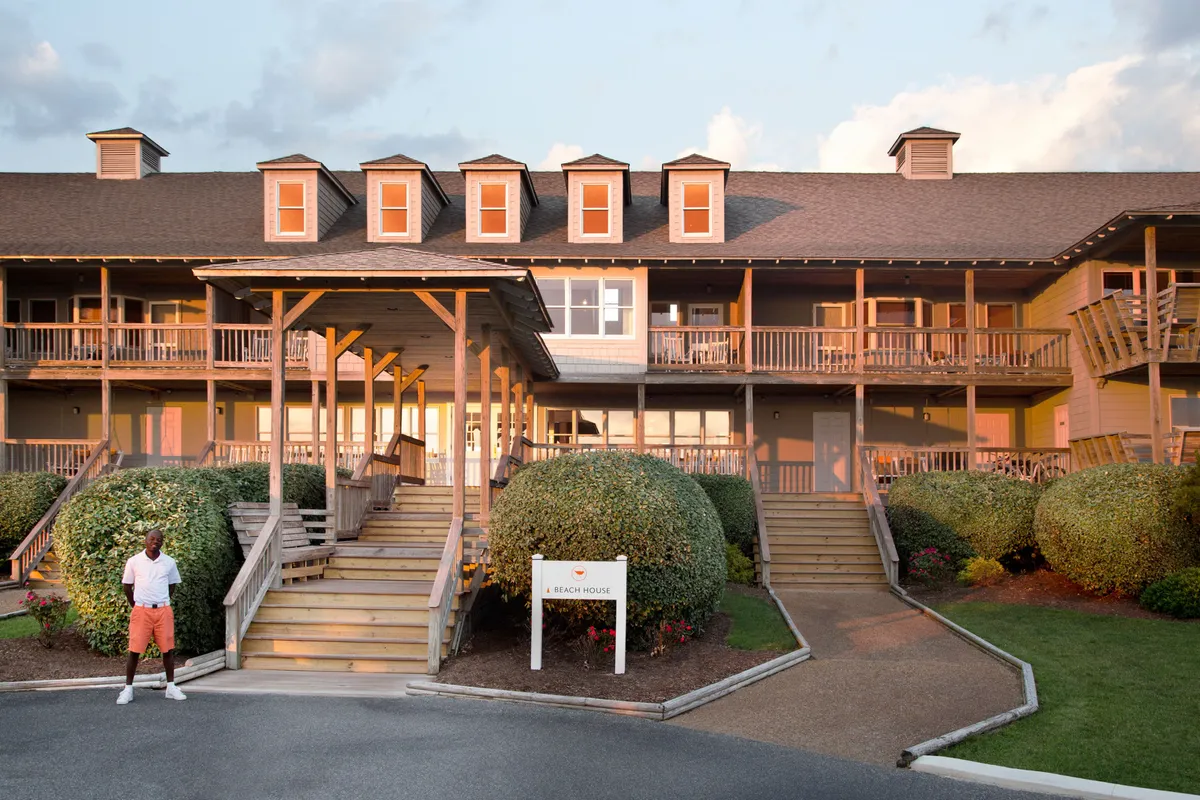 An inviting exterior shot featuring the entrance of 'A Beach House,' with wooden steps leading up to the porch, a sign indicating the name of the property, well-maintained landscaped bushes, and a person standing in front. The overall mood is cheerful and welcoming, suggesting the start or end of an outdoor adventure.