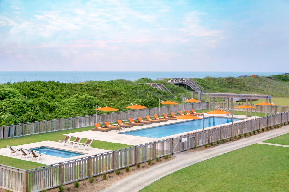 A serene resort swimming pool framed by lush greenery and orange umbrellas, with the ocean visible in the background. Lounge chairs encircle the sparkling pool, offering inviting spots for sunbathing, relaxing, and socializing in a casual yet picturesque setting. The environment exudes a sense of vacation relaxation and vibrant community.