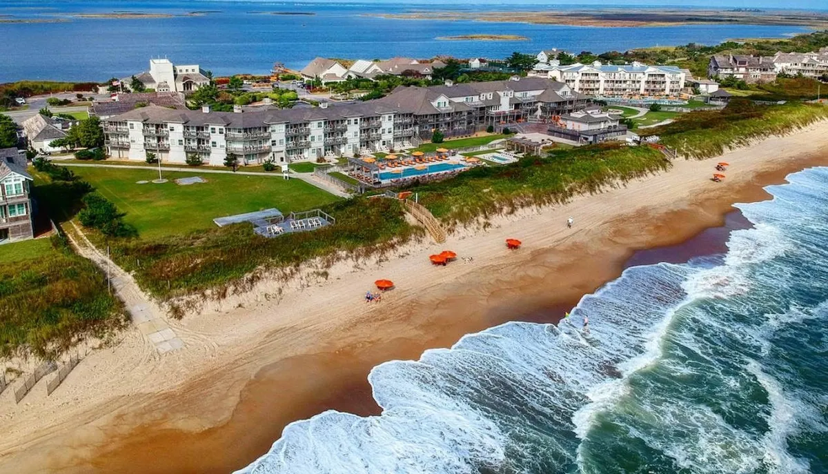 A vibrant exterior shot capturing the beachfront hotel with its sparkling swimming pool in the foreground, beach umbrellas set up for lounging, and ocean waves rolling gently beyond. There’s a clear sense of vacation leisure, with spaces for both poolside fun and easy access to the sandy shore.