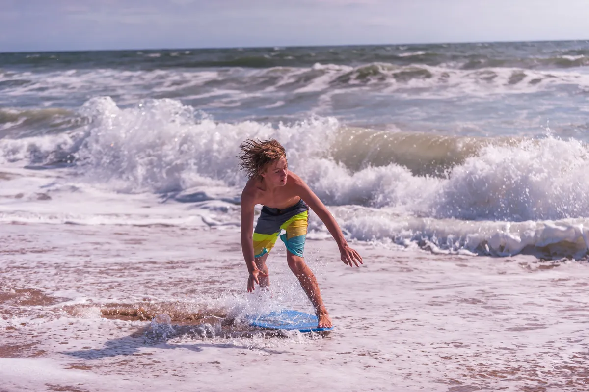 An action-filled beach scene featuring a boy riding a boogie board as waves crash around him beneath a blue sky, capturing the energy and joy of playing in the water.