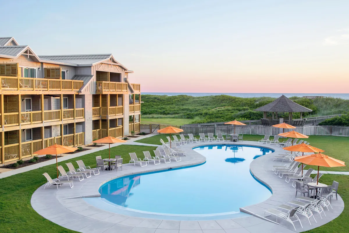A beautifully landscaped swimming pool area surrounded by patio umbrellas and lounge chairs, with the elegant hotel facade as a backdrop. The mood is both cheerful and serene, hinting at leisurely swims, relaxing poolside, or sipping drinks while enjoying expansive views.