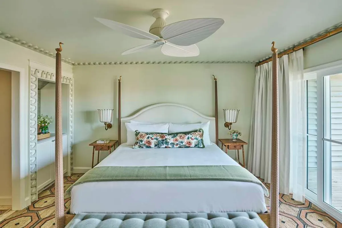 The image showcases an inviting studio interior with a plush four-poster king bed adorned with crisp linens and a floral accent pillow. A mirrored dresser reflects the soft natural light pouring in, while a ceiling fan suggests breezy comfort. The space feels tranquil and elegantly understated, ideal for relaxation or enjoying a morning coffee in bed.